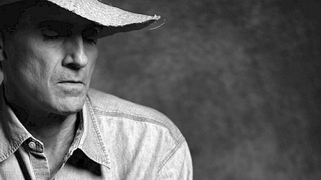 A person wearing a cowboy hat and denim shirt holds a guitar, with a serious expression against a textured background.