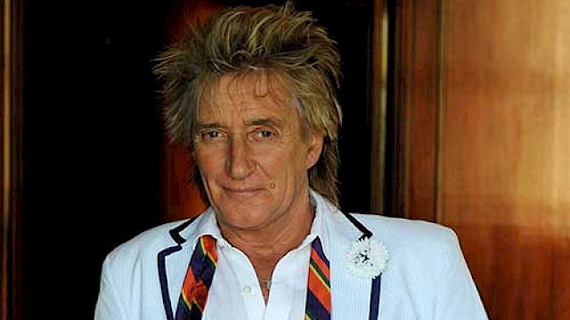 A person in a white suit jacket and colorful tie stands in front of a wooden background, wearing a pocket square and a small boutonniere.