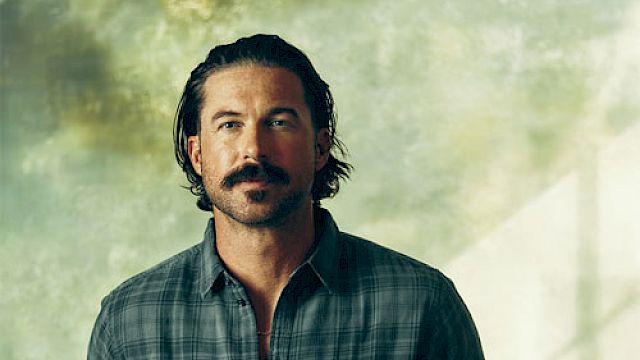 A man with a mustache and beard wearing a plaid shirt stands against a textured background, hands in pockets.