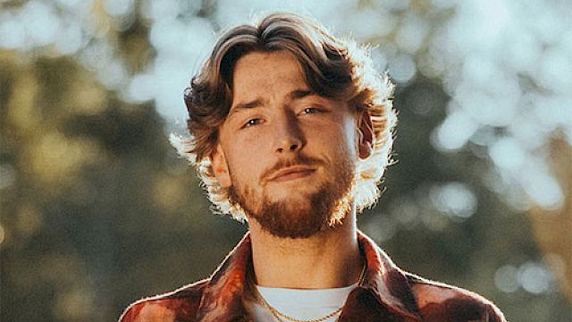 A person with a beard wearing a patterned shirt and necklaces stands outdoors with a blurred background of trees and sunlight.