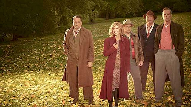 A group of people in vintage attire standing on a leaf-covered lawn with trees in the background.