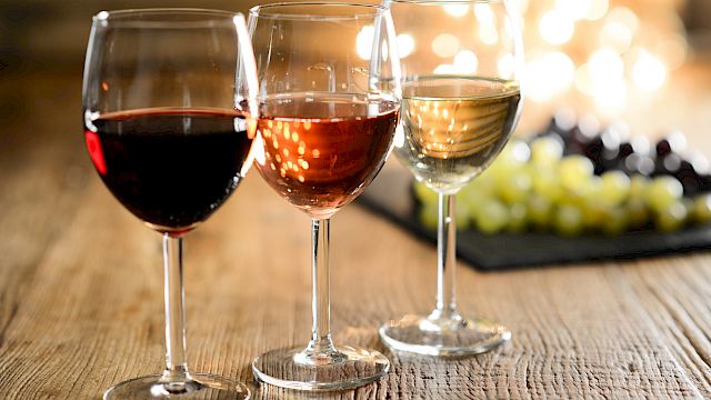 Three wine glasses filled with red, rosé, and white wine are on a wooden table, with grapes out of focus in the background.