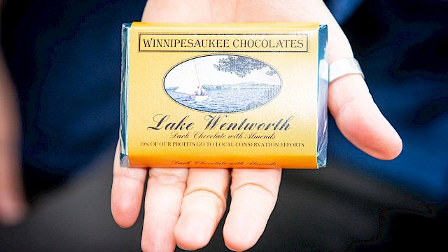 A hand holds a yellow bar of "Winnipeasaukee Chocolates," specifically "Lake Wentworth" with a sailboat illustration on the wrapper.