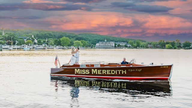 A couple stands on the "Miss Meredith" boat on a lake during sunset, with a picturesque landscape in the background.