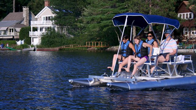 A family on a pedal boat enjoys a ride on a lake with waterfront houses and lush greenery in the background, all wearing life vests.