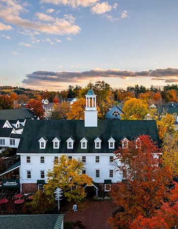 A picturesque aerial view of a quaint town with autumn foliage, charming buildings, and a distinctive central tower under a partly cloudy sky.