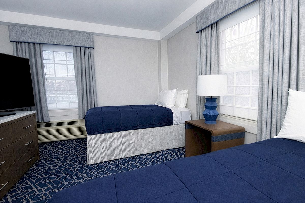 Two double beds with blue comforters in a bright hotel room, a dresser with a TV, and a nightstand with a blue lamp between two windows.