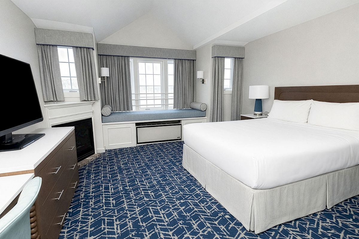 A well-lit hotel room with a king bed, blue patterned carpet, a desk, TV, fireplace area, and a window seat under two large windows.