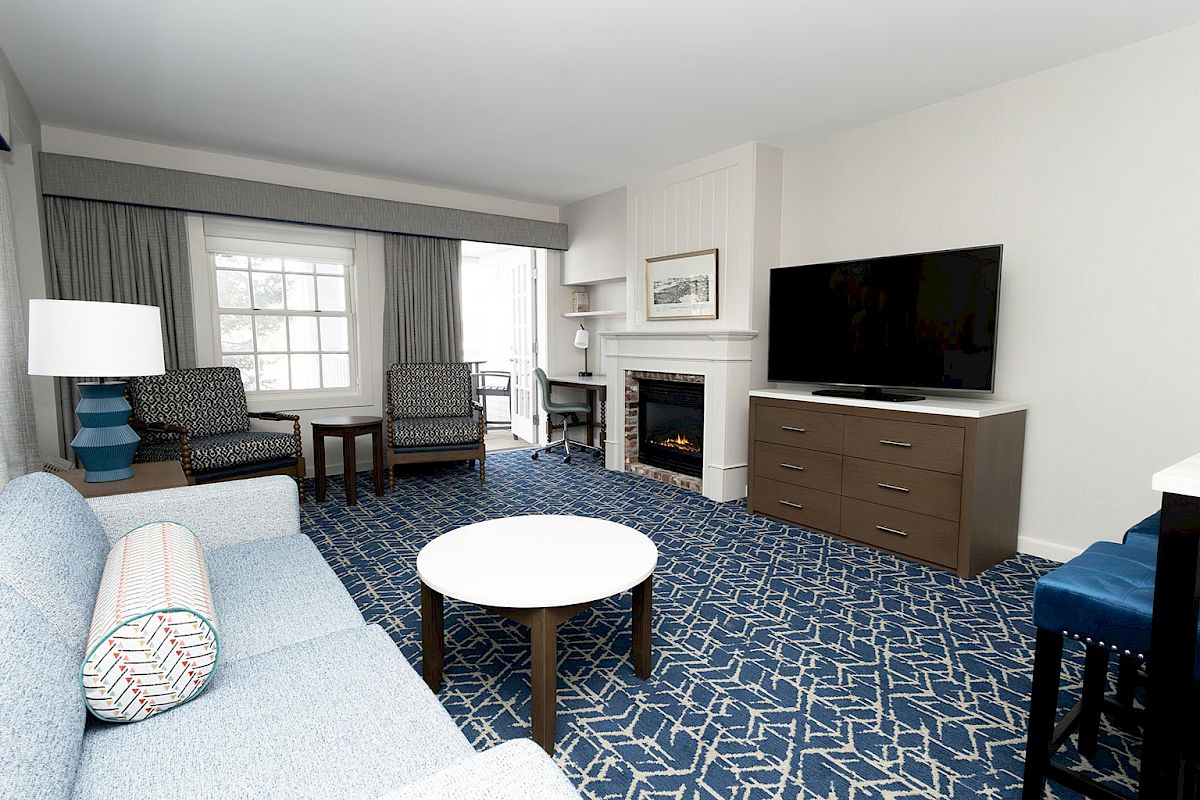 A hotel suite living area with a blue patterned carpet, sofa, two armchairs, coffee table, fireplace, flat-screen TV, dresser, and large window.
