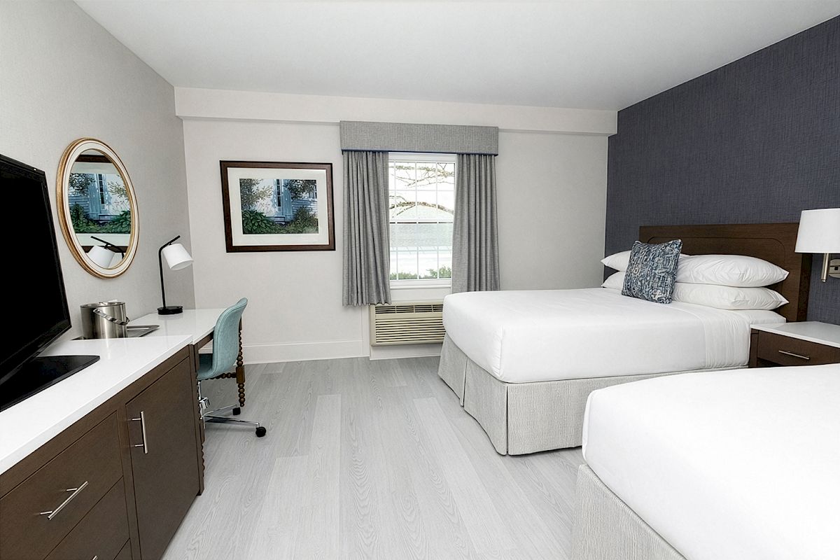 A bright hotel room with two queen beds, a dark accent wall, a desk and chair, a wall mirror, and a window with curtains.
