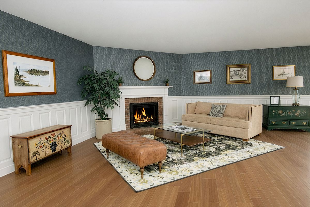 A cozy living room with a lit fireplace, beige sofa, patterned area rug, tufted leather ottoman, wooden end table, potted plant, and blue wallpapered walls.