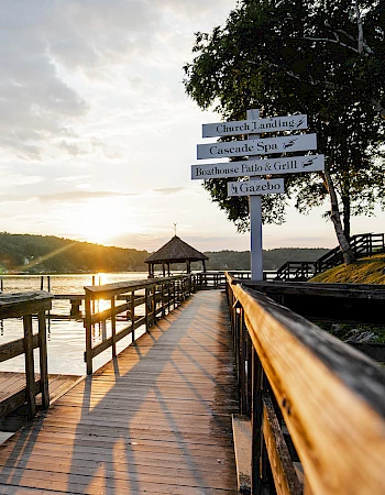 A wooden pier extends into a calm lake at sunset, with signposts and a small gazebo to the right, casting long shadows along the boardwalk.