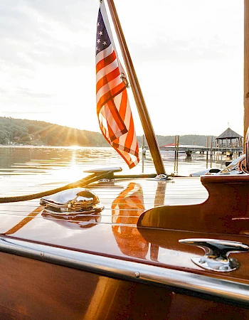 A polished wooden boat by the water at sunset, American flag fluttering, calm lake, and dock in the background.